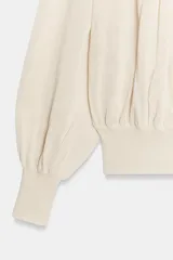 Blusa de punto color beige con cuello barco, manga larga abullonada con puños ajustados y detalles fruncidos en la parte delantera.