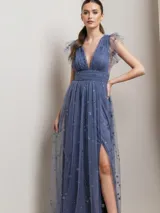 Vestido largo de fiesta en color azul grisáceo, confeccionado en tul con detalles de estrellas brillantes bordadas. Presenta un escote en V profundo, mangas cortas con volados de tul, cintura entallada y una falda larga con tajo lateral.