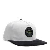 Gorro tipo snapback con corona gris y visera plana de textura aterciopelada en color negro. Presenta un parche circular negro en el frente con detalles gráficos y texto en color verde.