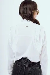 Camisa blanca de manga larga con cuello alto con volados, detalles de alforzas y pliegues en el frente y cierre frontal con botones ocultos.