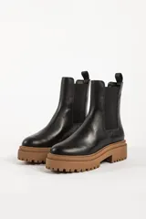 Bota tipo chelsea con suela track de goma en color marrón y capellada de cuero negro. Presenta elásticos laterales y tirador trasero.