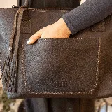 Cartera tipo tote de cuero marrón con costuras artesanales visibles, bolsillo frontal con el logo grabado, asas de hombro y detalles de flecos colgantes.