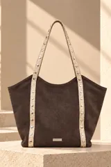 Cartera tipo tote de color marrón con cuerpo de gamuza y asas largas en tono tostado con textura de reptil y tachas metálicas plateadas. Cuenta con placa metálica con el logo de la marca en el frente.