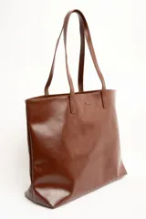 Bolso tipo tote bag color marrón, confeccionado en cuero sintético, con doble asa superior y detalle de placa metálica dorada con logo en la parte frontal.