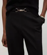 Pantalón de vestir negro confeccionado en malla punto roma, de corte largo y ligeramente afunilado. Presenta cintura alta con un adorno metálico decorativo en el frente y costuras verticales que estilizan la silueta.