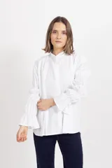 Camisa blanca de corte recto con cuello mao y cierre frontal oculto. Destaca por sus mangas largas con múltiples frunces que generan volumen y terminan en puños acampanados.