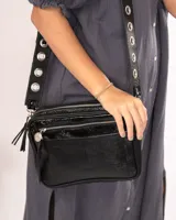 Bandolera negra de charol con doble compartimento, bolsillo frontal, correa regulable con ojalillos y detalles metálicos plateados.