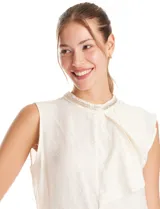 Blusa sin mangas color blanco, confeccionada en crepe con acabado satinado. Presenta un cuello alto decorado con mostacillas y una sobrecapa drapeada en el frente. La espalda cuenta con una abertura tipo gota y cierre de botón.