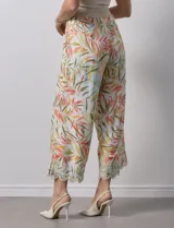 Pantalón capri de lino color crudo con estampado de hojas en tonos verdes, amarillos y naranjas. Presenta bolsillos laterales y detalle de puntilla en el ruedo.
