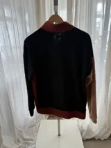 Buzo de corderito con diseño tricolor, que combina cuerpo en tono tostado, una manga negra y otra en color ladrillo. Cuenta con cuello alto con botones frontales y bolsillo tipo canguro.