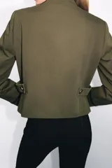 Blazer de estilo militar confeccionado en algodón, con cuello subido y manga larga. Presenta un diseño estructurado con detalle de alamares negros a contraste en el frente, botones metálicos decorativos y trabillas laterales. Cuenta con cierre frontal oculto y forro interior.