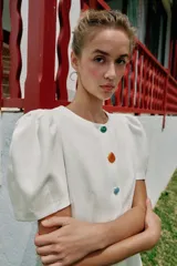 Vestido corto de color blanco con cuello redondo y mangas cortas abullonadas. Presenta un cierre frontal con botones decorativos de colores.