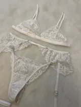Conjunto de lencería de encaje blanco compuesto por un bralette triangular, una tanga y un portaligas con volados en la cadera y ligas integradas.