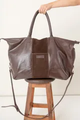 Bolso grande de cuero vacuno color marrón, con cierre superior, doble asa de mano y correa bandolera regulable y desmontable.