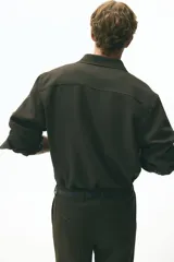 Camisa de manga larga color marrón oscuro, confeccionada en sarga suave, con cuello americano y tapeta clásica de botones. Presenta canesú en la espalda y bajo redondeado.