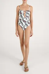 Malla enteriza de color blanco con estampado paisley en tonos azules, marrones y rojos. Presenta diseño bandeau con tirantes finos ajustables, escote con aberturas circulares unidas por argollas metálicas doradas.