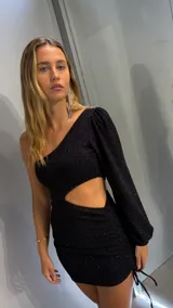 Vestido corto negro de tejido brillante con un solo hombro descubierto (asimetrico) y manga larga abullonada. Presenta un corte tipo cut-out en la zona del abdomen y frunces ajustables con cordones en la falda.