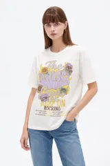 Remera blanca de manga corta, con cuello redondo y diseño holgado. Presenta un estampado frontal de estilo vintage con la inscripción "The Endless Tour Keep on Rocking" acompañada de ilustraciones de girasoles y otros detalles gráficos.