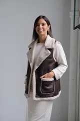 Chaleco de cuero sintético color chocolate con interior y bordes de corderito beige. Presenta solapas amplias, dos bolsillos frontales con detalle de corderito y corte recto.