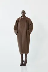 Abrigo largo tipo sobretodo de color marrón, confeccionado en una mezcla de poliéster y lana. Presenta mangas ranglán, pinzas frontales que aportan estructura y un cuello alto tipo tortuga con cierre cruzado mediante un botón.