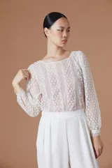 Blusa de encaje blanco con mangas largas abullonadas y puños con detalle de terminación desflecada. Presenta un diseño de cuello redondo y corte recto.