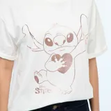 Camiseta de cuello redondo y manga corta, color blanco hueso, con estampado central metalizado en tono oro rosa del personaje Stitch sosteniendo un corazón.