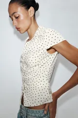 Camisa de cuello solapa y escote en pico, con mangas cortas y detalle de frunces centrales. Presenta cierre frontal con botones y estampado de lunares negros sobre fondo blanco.