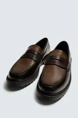 Mocasín de cuero marrón oscuro con antifaz en tono camel. Presenta horma redonda y suela track gruesa de goma negra.
