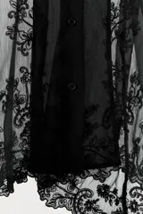 Blusa semitransparente negra de cuello solapa y manga larga, con cierre frontal de botones y detalles de bordados tonales en el ruedo y los puños.
