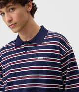 Remera polo de algodón con diseño a rayas horizontales en azul marino, rojo y blanco, corte boxy amplio, mangas cortas y cuello polo con botones. Presenta un pequeño bordado en el pecho.