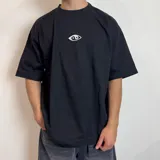 Remera de corte oversize confeccionada en algodón pesado, color negro. Presenta un parche rectangular cosido en la espalda con un diseño gráfico artístico en blanco y negro que muestra una mano alcanzando un círculo oscuro sobre un fondo de líneas horizontales.