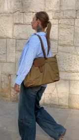Cartera tipo tote bag color marrón con detalles en cuero sintético tostado, solapa frontal con tira decorativa, doble asa de mano y refuerzos en las esquinas inferiores.