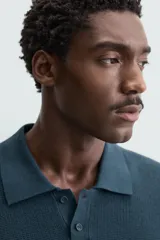 Polo de manga corta color verde azulado oscuro, confeccionado en tejido de punto con textura. Presenta cuello solapa y cierre frontal de botonadura.