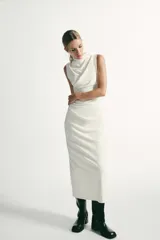 Vestido midi de color negro, sin mangas, con cuello subido y detalle de tejido drapeado en el lateral. Cuenta con cierre de cremallera oculta en la espalda.