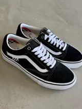 Championes Vans Skate Old Skool, color negro con franja lateral blanca, cordones blancos y suela blanca con línea negra. Presentan un detalle de bandera a cuadros en el lateral.