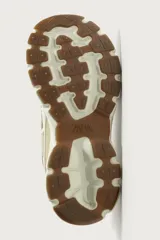 Zapatilla deportiva tipo running con diseño de suela voluminosa e irregular. Presenta una combinación de piezas en tonos beige y crema, con paneles de malla y cordones jaspeados.