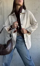 Chaqueta tipo sobrecamisa color beige, con cierre frontal de botones metálicos, cuello clásico y bolsillos frontales. Presenta un diseño de corte recto y mangas largas con puños abotonados.