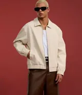 Campera masculina de corte boxy en color beige, con cuello inglés, cierre frontal de cremallera, mangas largas con puños elásticos y ruedo con elástico. Presenta bolsillos laterales y detalles de recortes en el diseño.
