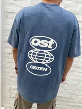Remera oversize de algodón con efecto acid wash en tono azul oscuro, con estampa blanca en la espalda que incluye los textos "Ost" y "Osten" enmarcados en óvalos, y un diseño de globo terráqueo en el centro.