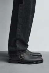 Botín de cuero negro con punta cuadrada, caña corta y cierre lateral con cremallera. Presenta una correa con hebilla metálica alrededor del tobillo.