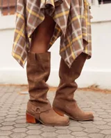 Botas de caña alta confeccionadas en cuero gamuzado color marrón. Presentan un diseño de estilo western con puntera cuadrada, taco de madera y una tira decorativa con estribo metálico desmontable en la zona del tobillo.
