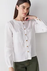 Blusa de lino color blanco, con cuello redondo, cierre frontal con botones de madera y dos bolsillos delanteros. Presenta mangas largas con puños abotonados y costuras a la vista en contraste.
