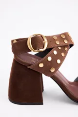 Sandalia de gamuza color marrón con taco cuadrado y punta abierta. Presenta tiras cruzadas en el tobillo con hebilla metálica dorada y detalle de tachas circulares doradas en las tiras y en la parte frontal.