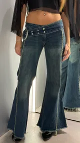 Jean de tiro bajo con diseño acampanado y costuras laterales expuestas con acabado deshilachado. Presenta una cintura ancha con detalle de hebilla metálica grande y ojales decorativos.