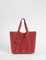 Cartera tipo shopper de gran tamaño confeccionada en material efecto gamuza color rojo. Presenta correas reforzadas decoradas con tachas metálicas plateadas y un dije colgante en forma de corazón con detalles de tachas. Cuenta con una placa metálica pequeña con logo en la parte inferior frontal.