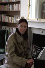 Trench coat largo color verde oliva/beige, con cuello solapa, manga larga ajustable con trabilla y botón, bolsillos delanteros y cierre frontal con botones. El forro interior visible en el cuello es de tela estampada a cuadros en tonos marrones.