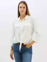 Camisa blanca de rayón con textura sutil, cuello con solapa, cierre frontal con botones y bolsillos simulados en el pecho. Presenta mangas 3/4 regulables con presilla y un diseño con nudo en el ruedo frontal.