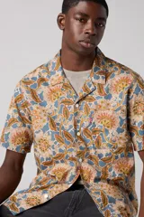 Camisa de manga corta con estampado floral en tonos beige, marrón y azul. Tiene cuello tipo camp y un bolsillo en el pecho.