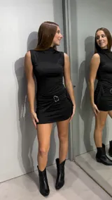 Vestido corto negro sin mangas, con cuello alto y diseño ajustado al cuerpo. Presenta un detalle de frunces en la cintura con una hebilla metálica decorativa.