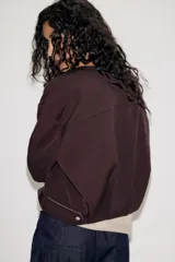 Chaqueta estilo bomber de color marrón oscuro, con cuello redondo, manga larga y cierre frontal con cremallera. Presenta bolsillos delanteros de vivo y un diseño con bajo fruncido.